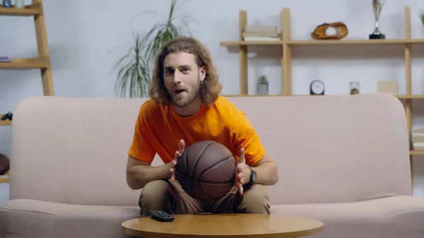 Begeisterter Sportfan mit Ball sitzt zu Hause auf der Couch und schaut Basketballspiel im Fernsehen — Stockfoto