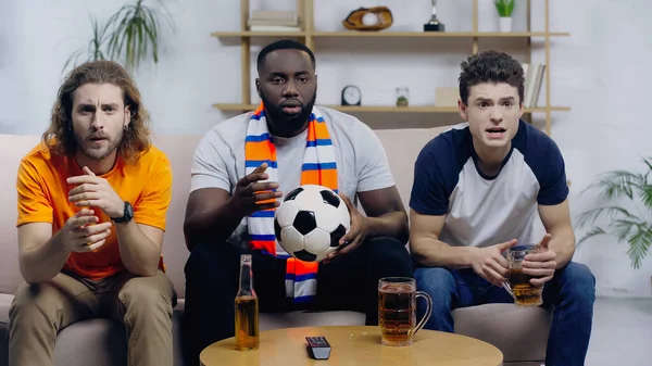 Worried multiethnic sport fans watching football championship near beer on coffee table — Stock Photo