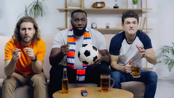 African american man with soccer ball holding clenched fist for luck while watching football match with worried friends — Stock Photo