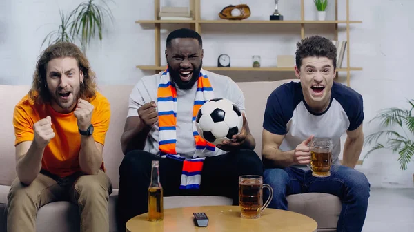 Overjoyed multicultural sport fans screaming and showing triumph gesture while watching football match — Fotografia de Stock