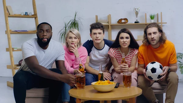 Tense multiethnic friends sitting on sofa near beer and chips while watching football match — Stock Photo