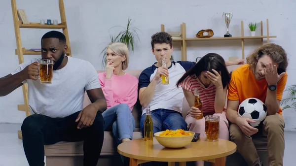 African american man drinking beer while watching football match with frustrated friends — Stock Photo