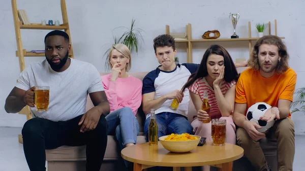 Upset multiethnic sport fans watching football match while sitting on sofa near beer and chips — Stock Photo