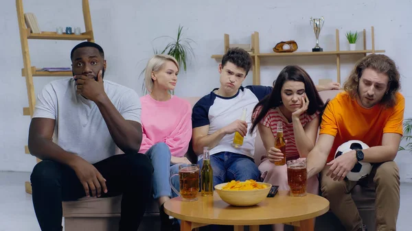 Unhappy multicultural sport fans watching football match on tv near chips and beer — Stock Photo