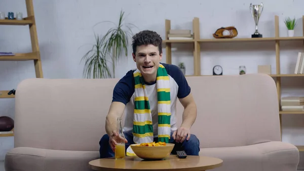 Fã de esporte emocionado em cachecol listrado assistindo competição no sofá perto de cerveja e batatas fritas — Fotografia de Stock