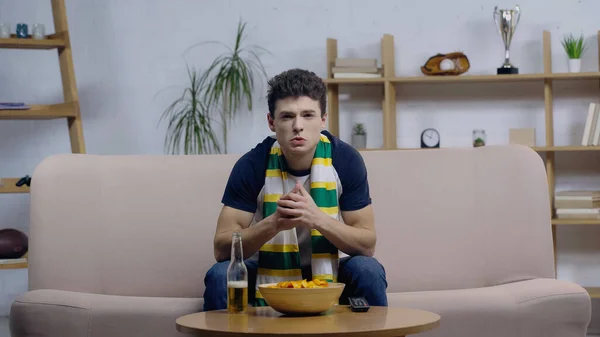 Worried young man watching sport match on tv at home near beer and chips — Stock Photo