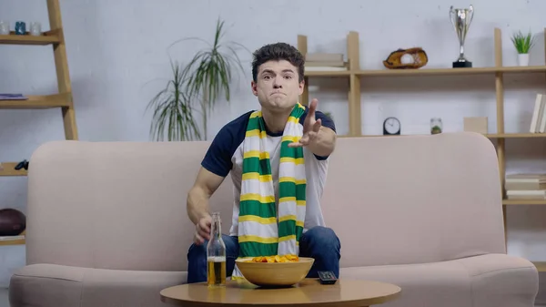 Thrilled man in striped scarf pointing with hand while watching sport competition on tv at home — Fotografia de Stock