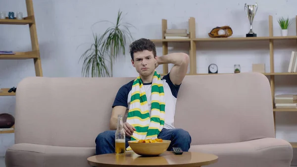 Discouraged sport fan sitting on sofa at home near beer and chips and watching championship — Fotografia de Stock