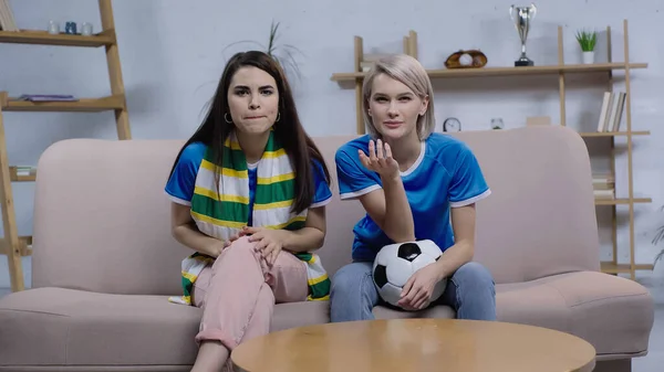 Mujeres tensas sentadas en el sofá con pelota de fútbol y bufanda rayada y viendo el partido de fútbol en la televisión - foto de stock