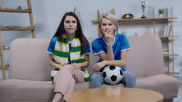 Mujeres tensas con bufanda a rayas y pelota de fútbol viendo el partido de fútbol en la televisión en casa - foto de stock