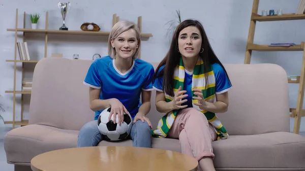 Thrilled women watching football championship while sitting on sofa with soccer ball — Stock Photo
