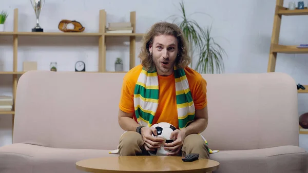 Изумленный фанат спорта в полосатом шарфе сидит на диване с мячом и смотрит чемпионат по футболу — стоковое фото