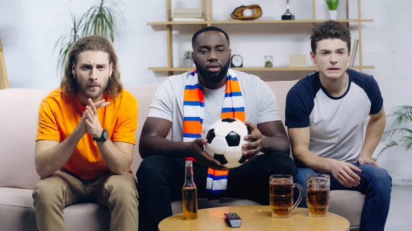 Concentrated multiethnic sport fans watching football game near beer on coffee table — Stock Photo