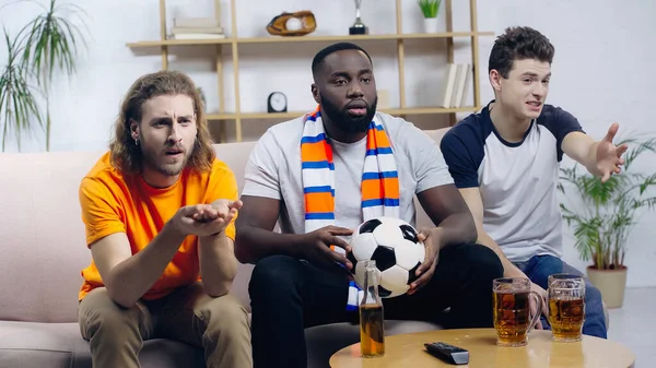 African american man with soccer ball watching football match near worried friends pointing with hands — Stock Photo