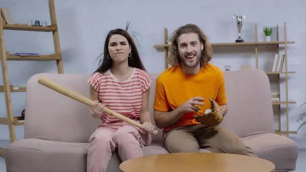 Displeased woman with baseball bat watching championship on tv near cheerful friend — Stockfoto