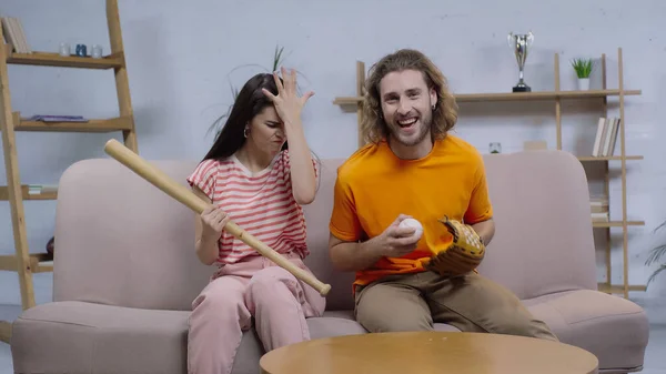 Cheerful man watching baseball game on tv while sitting near frustrated woman with baseball bat — Stock Photo