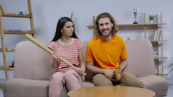 Angry woman with baseball bat looking at happy man watching match on tv at home — Fotografia de Stock