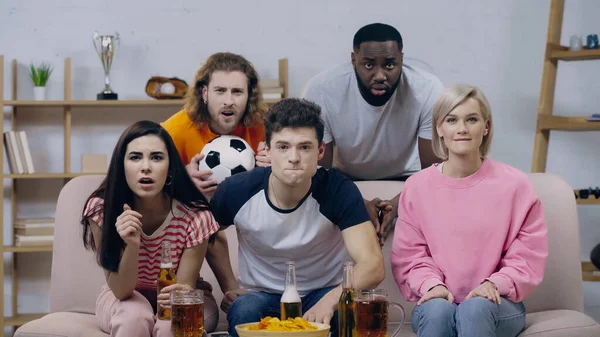 Excited man holding soccer ball while watching football match with worried multiethnic friends — Stock Photo