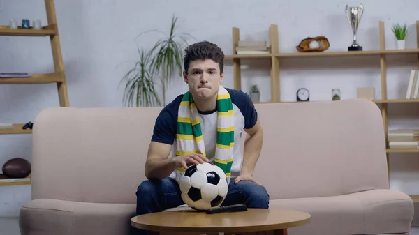 Nervöser Sportfan in gestreiftem Schal schaut Fußballspiel neben Ball auf Couchtisch — Stockfoto
