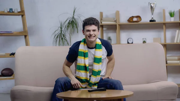 Cheerful young man sitting on sofa in striped scarf and watching sport competition — Fotografia de Stock