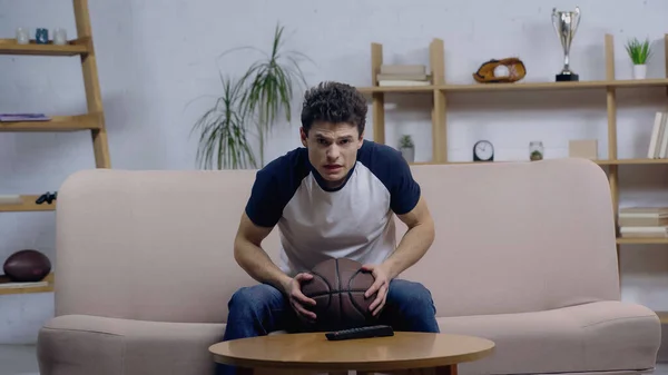 Нервовий фанат спорту дивиться баскетбольний матч на телевізорі, сидячи на дивані з м'ячем — стокове фото