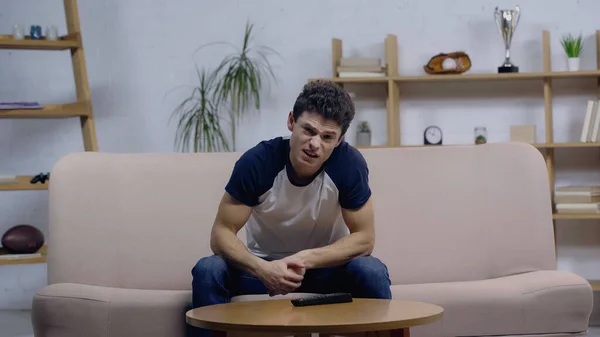 Jeune homme insatisfait de regarder la télévision tout en étant assis sur le canapé près de la table basse — Photo de stock