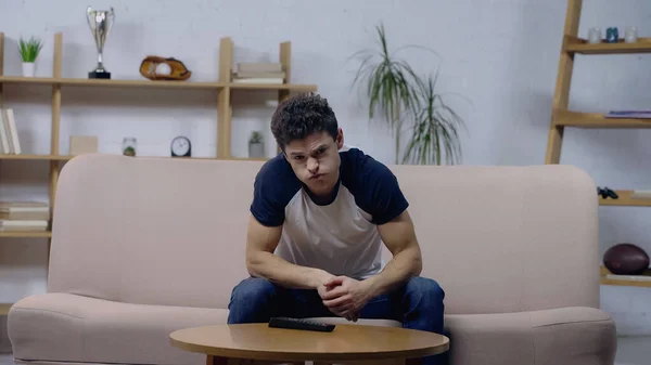 Aburrido joven hombre soplando mejillas mientras mira la televisión en el sofá en casa - foto de stock