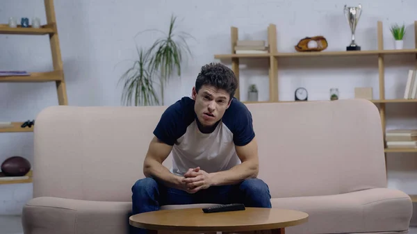 Unzufriedener junger Mann sitzt auf Couch neben Couchtisch und schaut Film im Fernsehen — Stockfoto