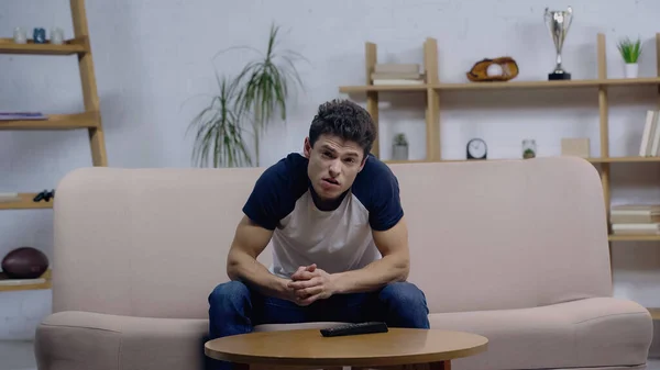 Displeased young man sitting on couch near coffee table and watching movie on tv — Stock Photo