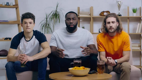 Concentrated multicultural friends watching sport championship near beer and chips at home — Stock Photo