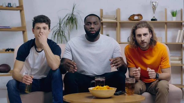 Worried man holding clenched fists for luck while watching sport match with multiethnic friends — Fotografia de Stock