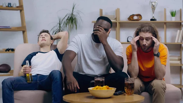 Frustrated multiethnic sport fans sitting near tv remote controller, beer and chips on coffee table — Stock Photo