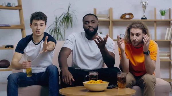 Frustrated multiethnic friends pointing with hands while watching sport game on tv at home — Stock Photo