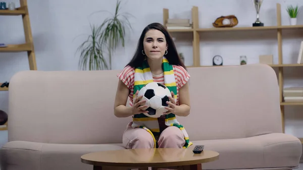 Femme concentrée en écharpe rayée tenant le ballon tout en regardant le match de football à la télévision à la maison — Photo de stock
