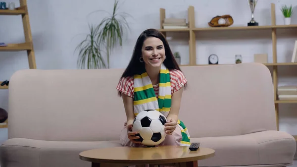 Mujer alegre en bufanda a rayas sentado en el sofá con pelota de fútbol y viendo el campeonato en la televisión - foto de stock