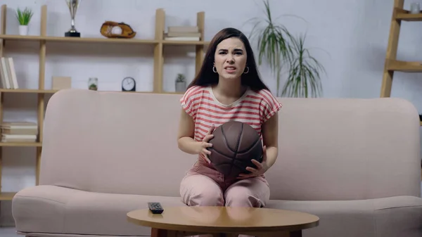 Worried sport fan woman holding ball while watching basketball match on home tv — Fotografia de Stock