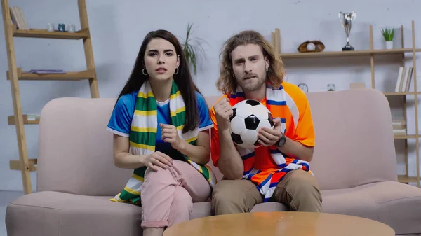 Концентрированные друзья в полосатых шарфах смотрят футбольный матч по телевизору дома — стоковое фото