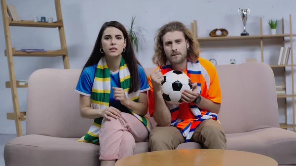 Concentrated friends in striped scarfs watching football match on tv at home — Stock Photo