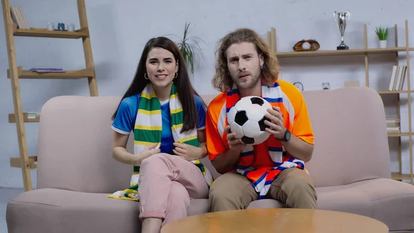 Worried sport fans in striped scarfs sitting on couch at home and watching football match — Stock Photo