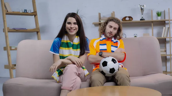 Cheerful woman in striped scarf sitting on couch near upset football fan — Fotografia de Stock