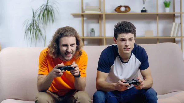 KYIV, UKRAINE - DECEMBER 22, 2021: excited man playing video game with focused friend on sofa at home — Foto stock