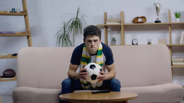 Нервный фанат спорта в полосатом шарфе держит мяч во время просмотра футбольного матча на домашнем телевидении — стоковое фото