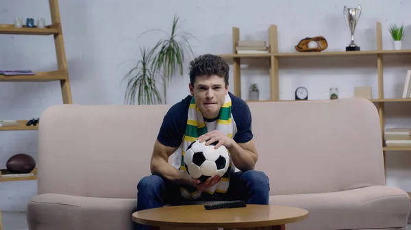 Tense football fan holding soccer ball while watching championship on couch at home — Fotografia de Stock