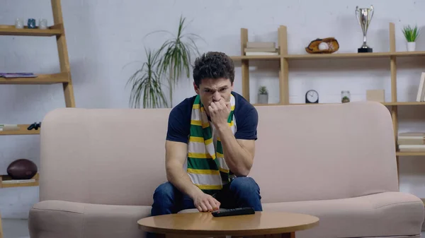 Frustrated sport fan in striped scarf sitting on sofa at home and watching match on tv — Stock Photo