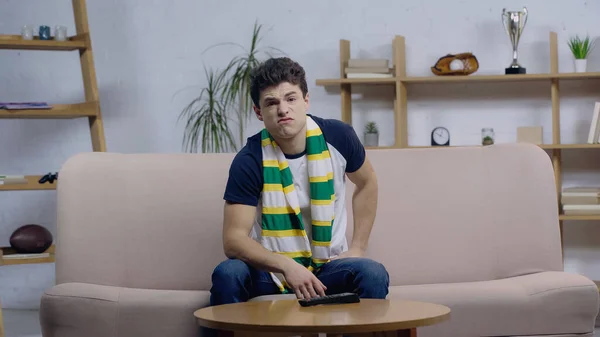 Frustrado joven fanático del deporte viendo el juego en la televisión mientras está sentado en el sofá en casa - foto de stock