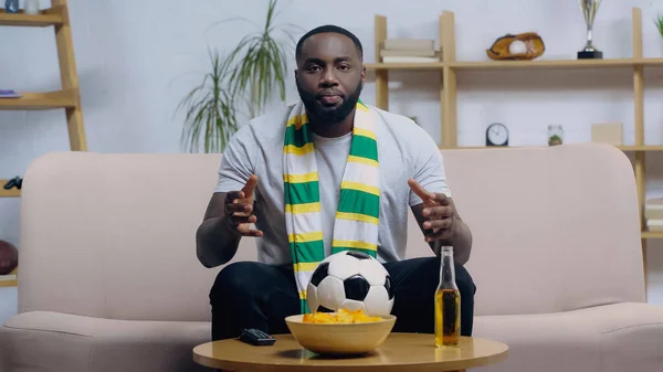 Besorgter afrikanischer Fußballfan gestikuliert, während er ein Spiel in der Nähe von Fußball, Bier und Chips im Fernsehen verfolgt — Stockfoto