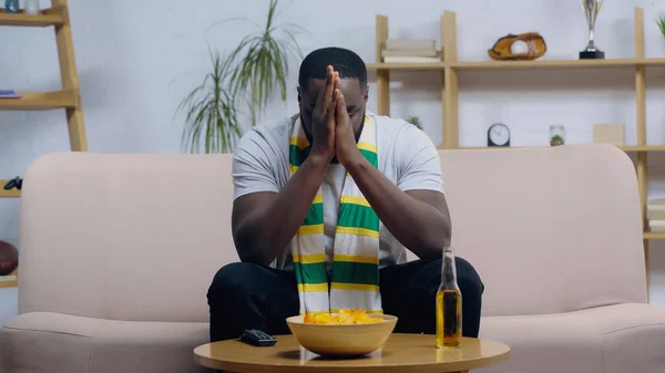 Torcedor de esporte afro-americano frustrado sentado perto da mesa de café com controle remoto tv, cerveja e batatas fritas — Fotografia de Stock