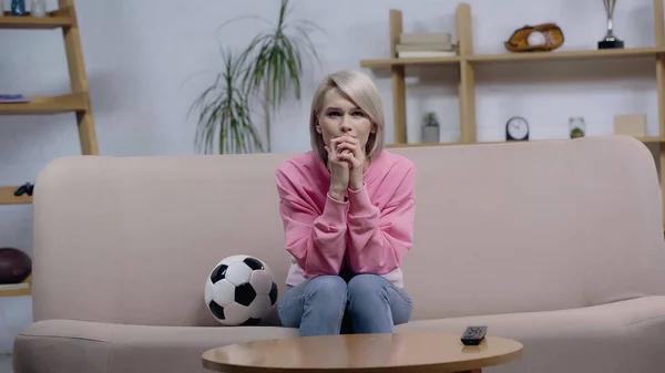 Nervioso deporte fan mujer cogido de la mano cerca de la cara mientras mira partido en sofá cerca de fútbol pelota - foto de stock