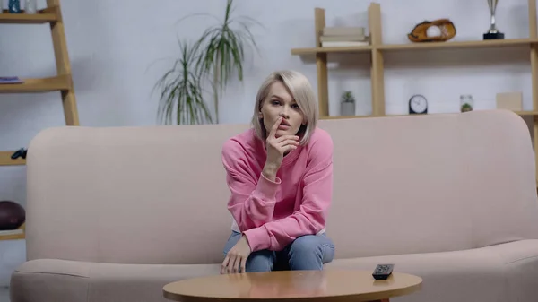 Femme blonde réfléchie assise sur le canapé à la maison et regardant un film à la télévision — Photo de stock