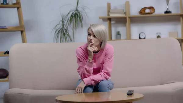 Femme blonde réfléchie assise sur le canapé près de la télécommande tv sur la table basse — Photo de stock